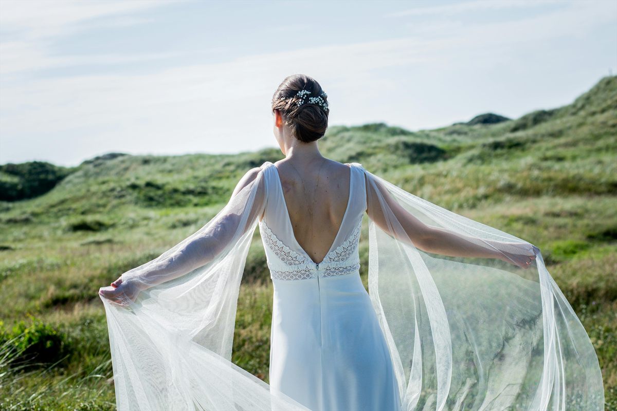 Braut von hinten in offenem Brautkleid mit fließendem Schleier auf einer Wiese