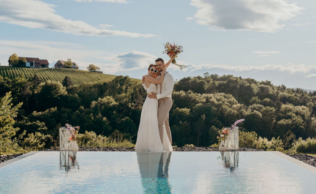 Brautpaar steht mit Blumenstrauß an einem Pool einer Hochzeitslocation mit blauem Himmel und Bergen.